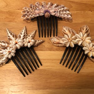 Peut inclure: Trois peignes à cheveux avec des dents en plastique noir, chacun décoré de coquillages naturels. Un peigne présente une disposition en éventail avec une perle violette au centre, tandis que les autres arborent des motifs en étoile et floraux.