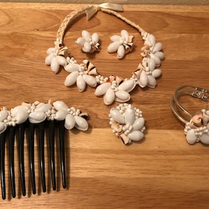 Puede incluir: Un conjunto de joyas de conchas blancas, que incluye un collar, pendientes, una pulsera y un peine para el cabello. El collar está hecho de conchas blancas enhebradas en un cordón de fibra natural. Los pendientes son pequeños y redondos, con un centro de concha blanca. La pulsera es una simple banda dorada con un diseño de concha blanca. El peine para el cabello es negro con detalles de concha blanca.