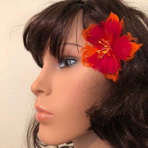 May include: A red and orange feather flower hair clip. The flower is made of feathers and has a yellow centre.