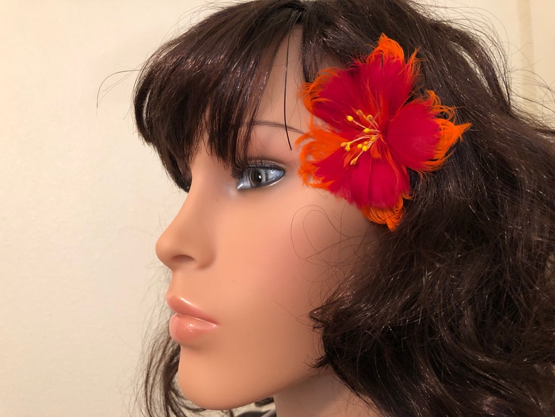 May include: A red and orange feather flower hair clip. The flower is made of feathers and has a yellow centre.