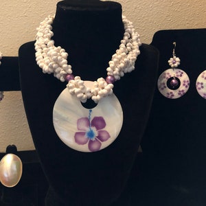 May include: A white shell necklace with a large shell pendant featuring a purple flower design. The necklace is paired with matching earrings and a ring, all with purple accents.