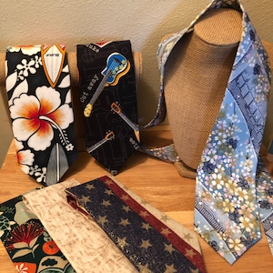 May include: A collection of neckties with various designs. One tie features a black background with a white hibiscus flower and a surfboard. Another has a blue ukulele graphic. Others have floral, star, and patterned designs. The ties are displayed on a wooden surface.