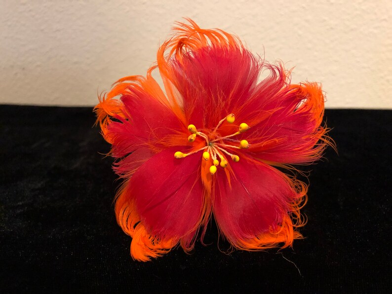 May include: A red and orange feather flower with a yellow centre. The flower is made of feathers and has a fluffy texture.