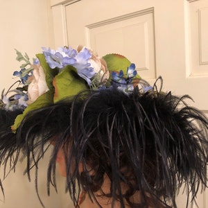 Pu&ograve; includere: Un cappello nero con piume e un grande gruppo di fiori bianchi e blu in cima.