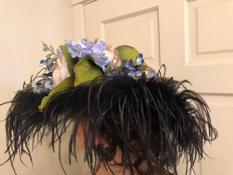 Pu&ograve; includere: Un cappello nero con piume e un grande gruppo di fiori bianchi e blu in cima.