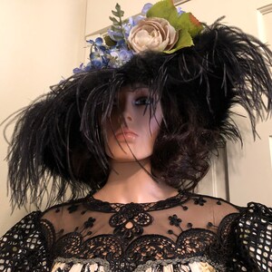 Pu&ograve; includere: Un cappello nero a tesa larga con piume e un ornamento floreale. Il cappello &egrave; adornato con fiori blu e bianchi e foglie verdi.
