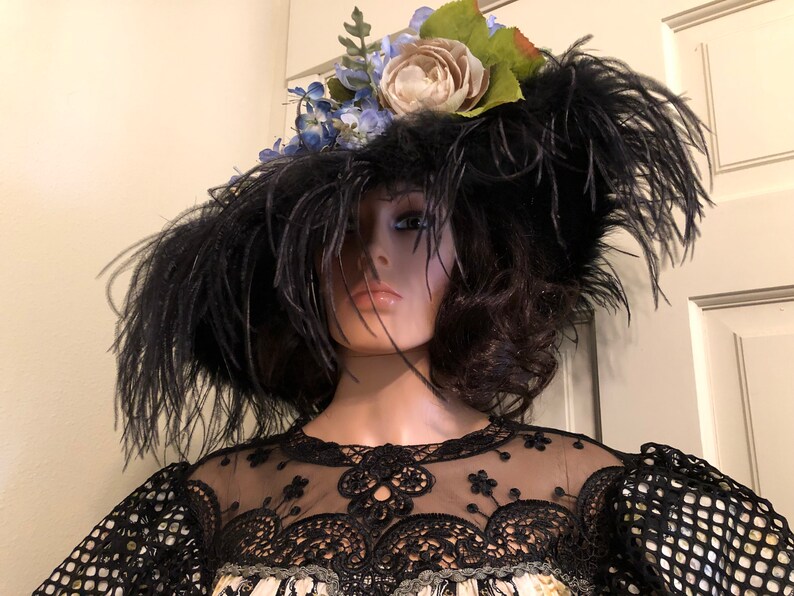 Pu&ograve; includere: Un cappello nero a tesa larga con piume e un ornamento floreale. Il cappello &egrave; adornato con fiori blu e bianchi e foglie verdi.