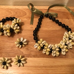 Può includere: Un set di gioielli composto da una collana, un braccialetto e orecchini. La collana e il braccialetto sono realizzati con perline nere e conchiglie di cauri. Gli orecchini sono realizzati con conchiglie di cauri e perline nere.