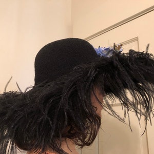 Pu&ograve; includere: Un cappello nero a tesa larga fatto di piume. Il cappello &egrave; decorato con fiori blu.