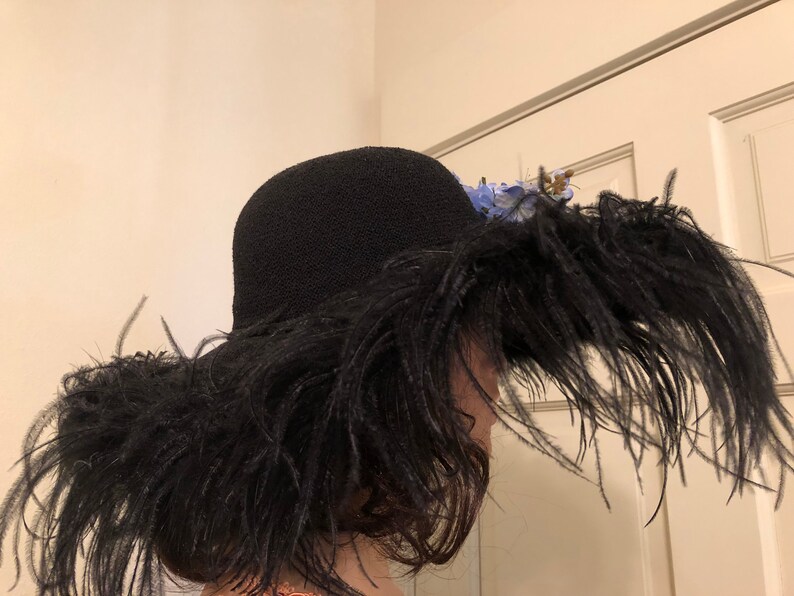 Pu&ograve; includere: Un cappello nero a tesa larga fatto di piume. Il cappello &egrave; decorato con fiori blu.