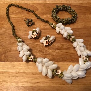 Könnte beinhalten: Ein Set aus weißem Muschelschmuck, bestehend aus einer Halskette, einem Armband und Ohrringen. Die Halskette und das Armband bestehen aus kleinen, weißen Muscheln, die mit grünen Perlen aufgereiht sind. Die Ohrringe bestehen aus größeren, weißen Muscheln mit einem grünen Perlenakzent.
