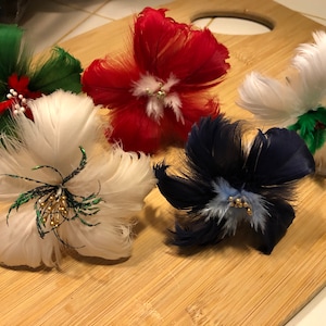 Puede incluir: Cinco adornos de flores de plumas en varios colores, incluyendo verde, rojo, blanco, azul y azul claro. Cada flor tiene un centro con cuentas doradas y detalles verdes.