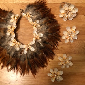 以下が含まれることがあります： 金色のアクセントが付いた白い貝殻の花のジュエリーセット。セットにはネックレス、ブレスレット、別々の花のピンが3つ含まれています。ネックレスは茶色の羽で囲まれています。