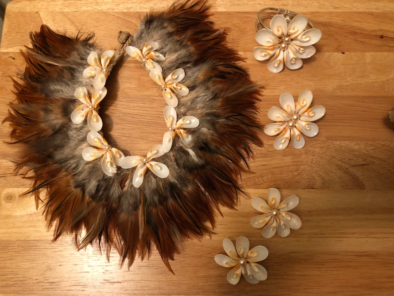 以下が含まれることがあります： 金色のアクセントが付いた白い貝殻の花のジュエリーセット。セットにはネックレス、ブレスレット、別々の花のピンが3つ含まれています。ネックレスは茶色の羽で囲まれています。