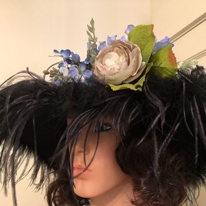 Pu&ograve; includere: Un cappello di piume nere con un grande fiore bianco e fiori blu in cima.