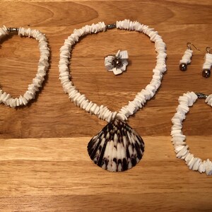 Pode incluir: Um conjunto de joias de concha branca, incluindo um colar com um grande pingente de concha, duas pulseiras e um par de brincos com contas marrons.