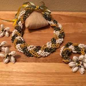 Könnte beinhalten: Ein Schmuckset aus Muscheln mit einer Halskette, einem Armband und Ohrringen. Die Halskette und das Armband bestehen aus weißen, grünen und gelben Muscheln. Die Ohrringe sind weiße Muschelblüten mit goldenen Akzenten.