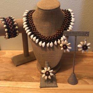 May include: A complete jewelry set featuring a necklace, bracelet, earrings, and ring. The necklace and bracelet are made of brown beads and white shells. The earrings and ring have a flower design with white shells and a brown center.