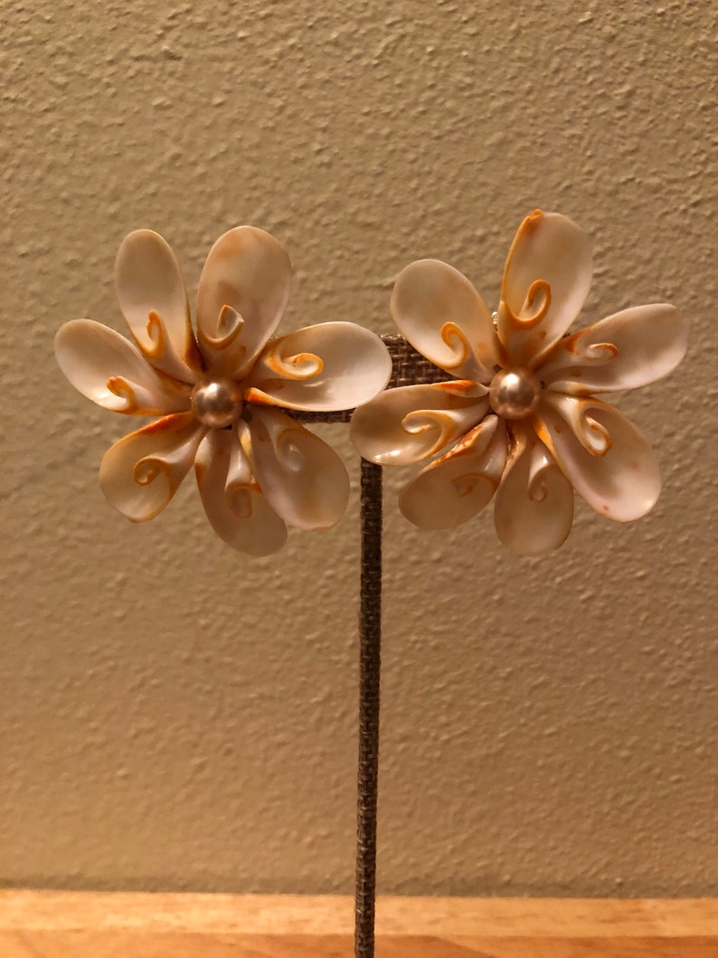 以下が含まれることがあります： 白い貝殻の花の形をしたイヤリングのペア。各花の中央にパールがあしらわれています。