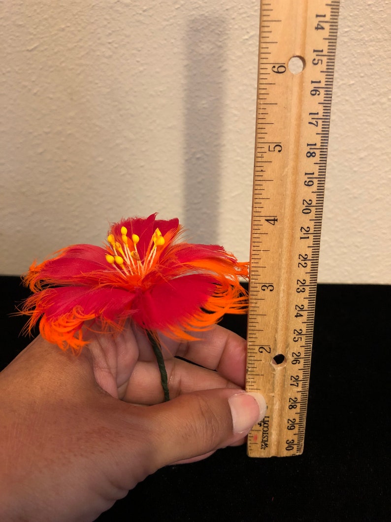 May include: A red and orange feather flower with a yellow centre. The flower is approximately 7.5 cm in diameter and is held in a hand next to a ruler.