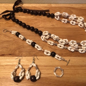 May include: A set of shell jewellery, including a necklace, bracelet, earrings, and a ring. The necklace and bracelet are made of white shells and brown wooden beads. The earrings are made of white shells and brown wooden beads. The ring is made of white shells.