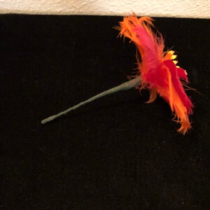 May include: A single orange and pink feather flower with a green wire stem. The flower is made of feathers and has a gold glitter accent.