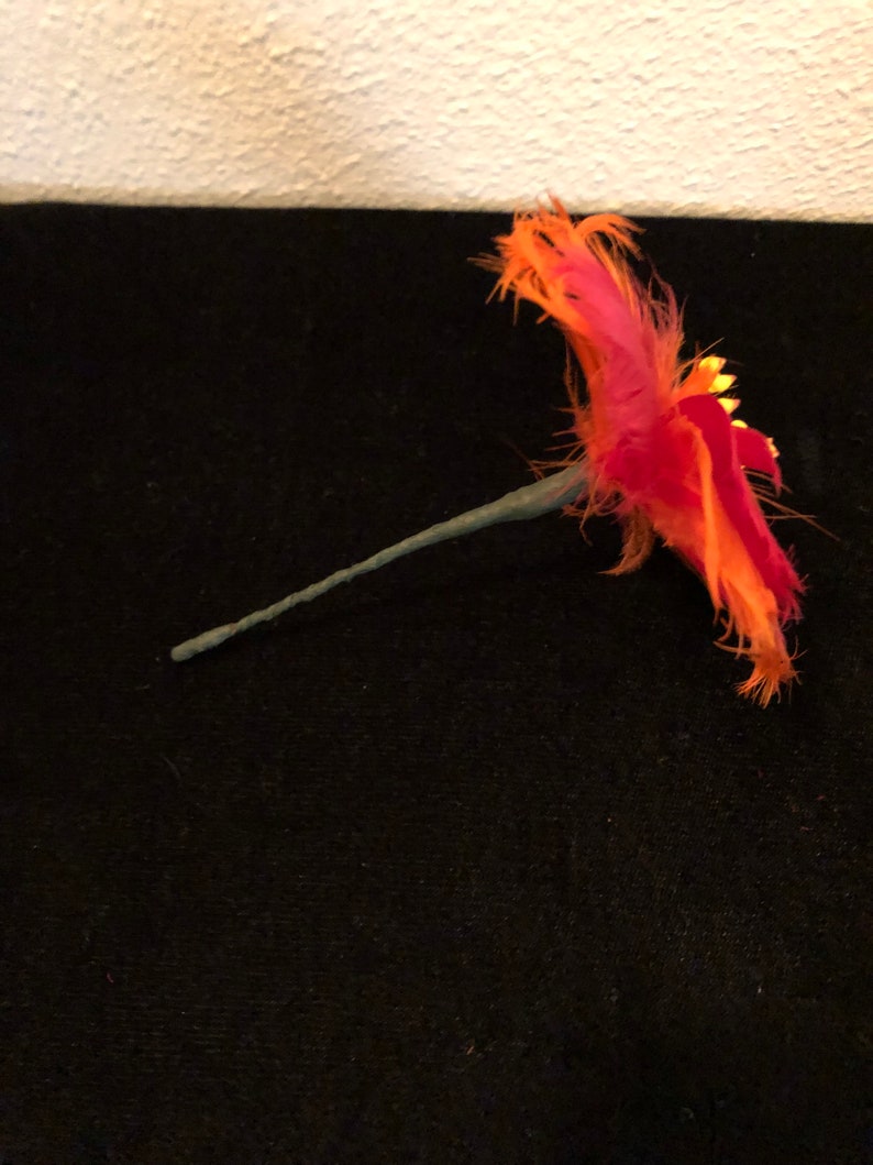 May include: A single orange and pink feather flower with a green wire stem. The flower is made of feathers and has a gold glitter accent.