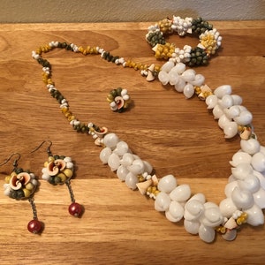 Könnte beinhalten: Ein Set aus Muschel-Schmuck, bestehend aus einer Halskette, einem Armband und Ohrringen. Die Halskette und das Armband sind aus weißen, gelben und grünen Muscheln gefertigt. Die Ohrringe sind aus weißen, gelben und grünen Muscheln mit einem roten Perlenakzent.