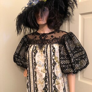 Pu&ograve; includere: Un abito bianco e nero con stampa floreale e dettagli in pizzo nero. L'abito ha maniche a sbuffo e una scollatura rotonda. La modella indossa un grande cappello nero con piume e fiori blu.