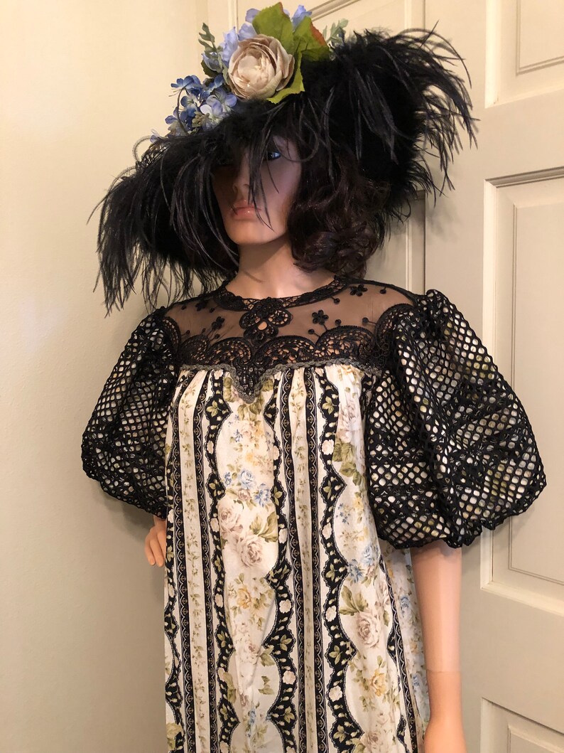 Pu&ograve; includere: Un abito bianco e nero con stampa floreale e dettagli in pizzo nero. L'abito ha maniche a sbuffo e una scollatura rotonda. La modella indossa un grande cappello nero con piume e fiori blu.