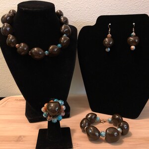 Op de afbeelding: Een set met een bruine zaadkraal ketting, armband en oorbellen met turquoise en koperen accenten. De ketting ligt op een zwarte fluwelen standaard, de armband ligt op een houten oppervlak en de oorbellen liggen op een zwarte fluwelen standaard.