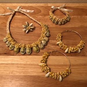 Könnte beinhalten: Ein Set aus vier Muschelkette in verschiedenen Größen. Die Ketten bestehen aus kleinen, weißen Muscheln und sind auf einer dünnen, weißen Schnur aufgefädelt. Die Ketten werden auf einer Holzoberfläche präsentiert.