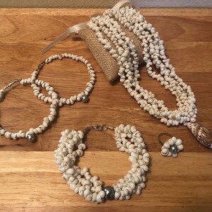 Könnte beinhalten: Ein Set aus weißem Muschel-Schmuck, bestehend aus einer Halskette, zwei Armbändern, einem Ring und einem Paar Ohrringen. Der Schmuck ist aus kleinen, weißen Muscheln gefertigt und hat einen silberfarbenen Akzent.