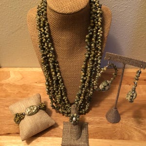 May include: A green and brown shell bead necklace, bracelet, ring, and earrings set. The necklace is multi-strand and the beads are irregular in shape. The set is displayed on a burlap background.
