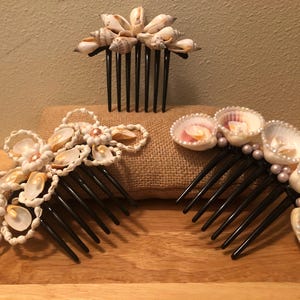 Könnte beinhalten: Drei schwarze Haarkämme, die mit natürlichen Muscheln verziert sind. Ein Kamm zeigt eine Ansammlung kleiner weißer Muscheln, ein anderer hat komplexe Muschelanordnungen mit weißen Schlaufen und der dritte zeigt Muscheln mit kleinen weißen Perlen.