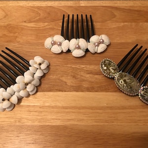 Puede incluir: Tres peines para el cabello decorados con conchas marinas. Los peines son de plástico negro con una fila de dientes. Un peine tiene conchas blancas dispuestas en forma de flor. Otro peine tiene conchas blancas con perlas rosas. El tercer peine tiene conchas verdes y marrones con pedrería.
