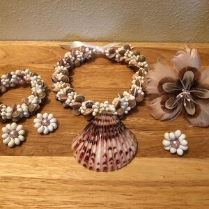 Könnte beinhalten: Ein Set aus Muschelschmuck, bestehend aus einer Halskette, einem Armband, Ohrringen und einer Muschelblume. Die Halskette und das Armband sind aus kleinen weißen und braunen Muscheln gefertigt. Die Ohrringe sind weiß mit einem rosa Zentrum. Die Blume ist braun mit weißen Federn.