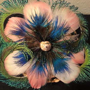 May include: A pink flower made of feathers with blue and green accents. The flower is centered around a small seashell.