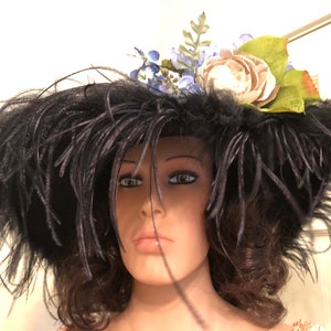 Pu&ograve; includere: Un grande cappello nero a tesa larga e piume nere. Il cappello &egrave; decorato con un fiore bianco e fiori blu.