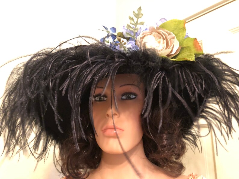 Pu&ograve; includere: Un grande cappello nero a tesa larga e piume nere. Il cappello &egrave; decorato con un fiore bianco e fiori blu.