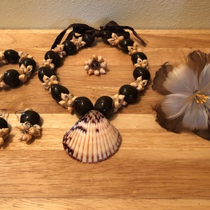 Puede incluir: Un conjunto de joyas de conchas marrones y blancas, que incluye un collar, una pulsera, pendientes y un colgante de concha. El conjunto también incluye una flor de plumas marrones y blancas.