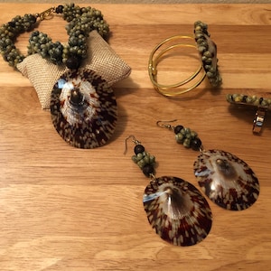Könnte beinhalten: Ein Schmuckset mit Halskette, Armband, Ring und Ohrringen. Halskette und Armband bestehen aus grünen Perlen und haben einen Muschelanhänger. Der Ring und die Ohrringe haben einen Muschelanhänger und grüne Perlen.