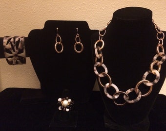 Op de afbeelding: Een set sieraden met een witte en zwarte gepatroneerde ketting, oorbellen, armband en ring. De ketting en oorbellen zijn gemaakt van goudkleurig metaal met witte en zwarte gepatroneerde schakels. De armband is een brede, witte en zwarte gepatroneerde manchet. De ring is een cluster van zwarte en witte kralen met een enkele witte parel in het midden.