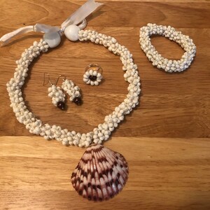 Könnte beinhalten: Ein Set aus weißem Muschel-Schmuck, bestehend aus einer Halskette, einem Armband, Ohrringen und einem Ring. Die Halskette hat einen großen Anhänger aus brauner Muschel.