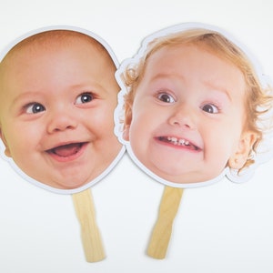 May include: Two white cardboard face masks with images of babies on them. The masks are attached to wooden sticks.