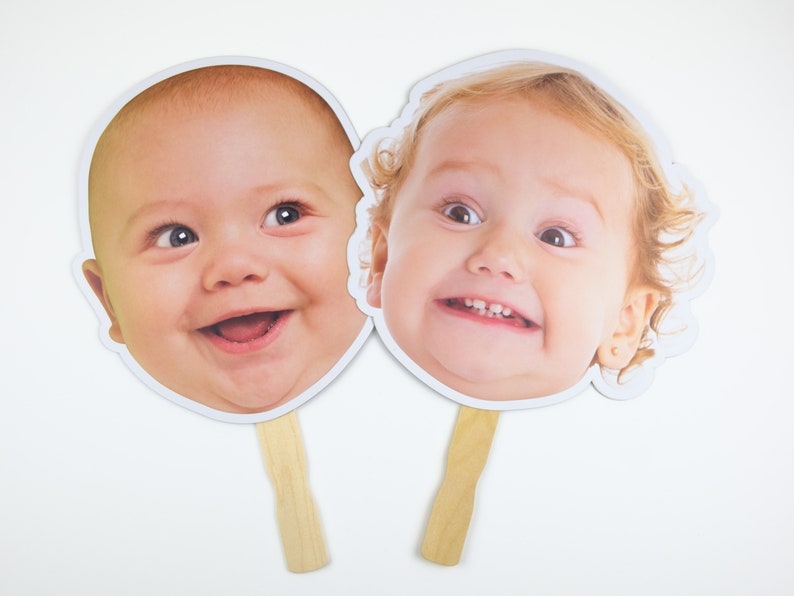 May include: Two white cardboard face masks with images of babies on them. The masks are attached to wooden sticks.