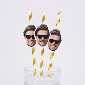 May include: Three decorative straws with a smiling man's face wearing sunglasses. The straws are white with gold stripes. The straws are in a clear glass filled with ice.