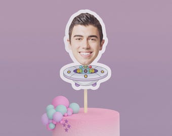 Könnte beinhalten: Ein Tortenaufsatz mit einem lächelnden Gesicht über einem Cartoon-Raumschiff-Design. Der Kuchen ist rosa mit einer Ansammlung von rosa, blauen und lila Luftballons und lila Sternenverzierungen. Der Hintergrund ist lavendelfarben.