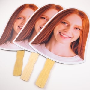 May include: Three white cardboard hand fans with a smiling woman's face printed on each. The fans have wooden sticks attached to the bottom.