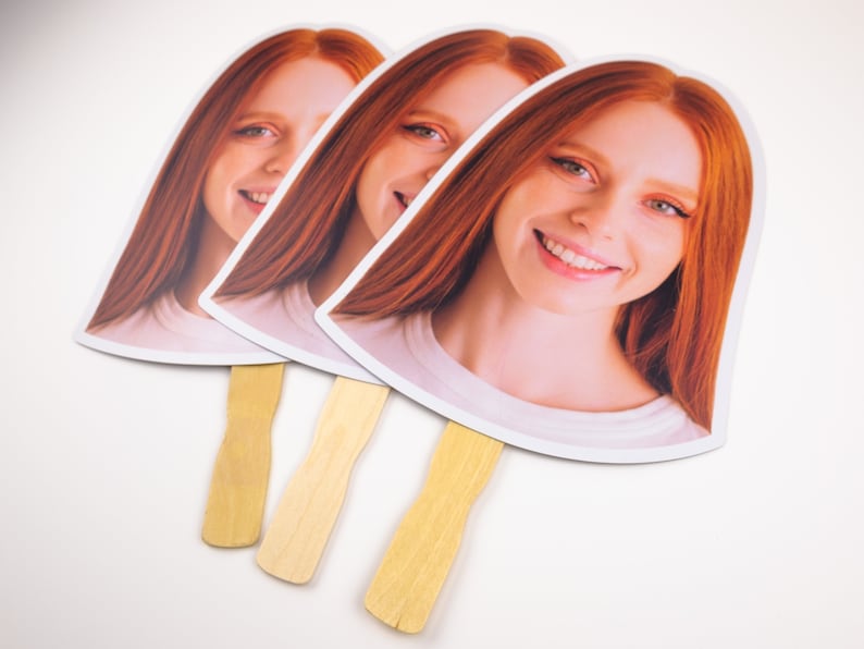 May include: Three white cardboard hand fans with a smiling woman's face printed on each. The fans have wooden sticks attached to the bottom.